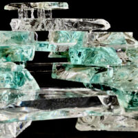 A small bench made of green glass with 4 legs of smokey shattered glass, sits atop a scattering of broken glass rocks