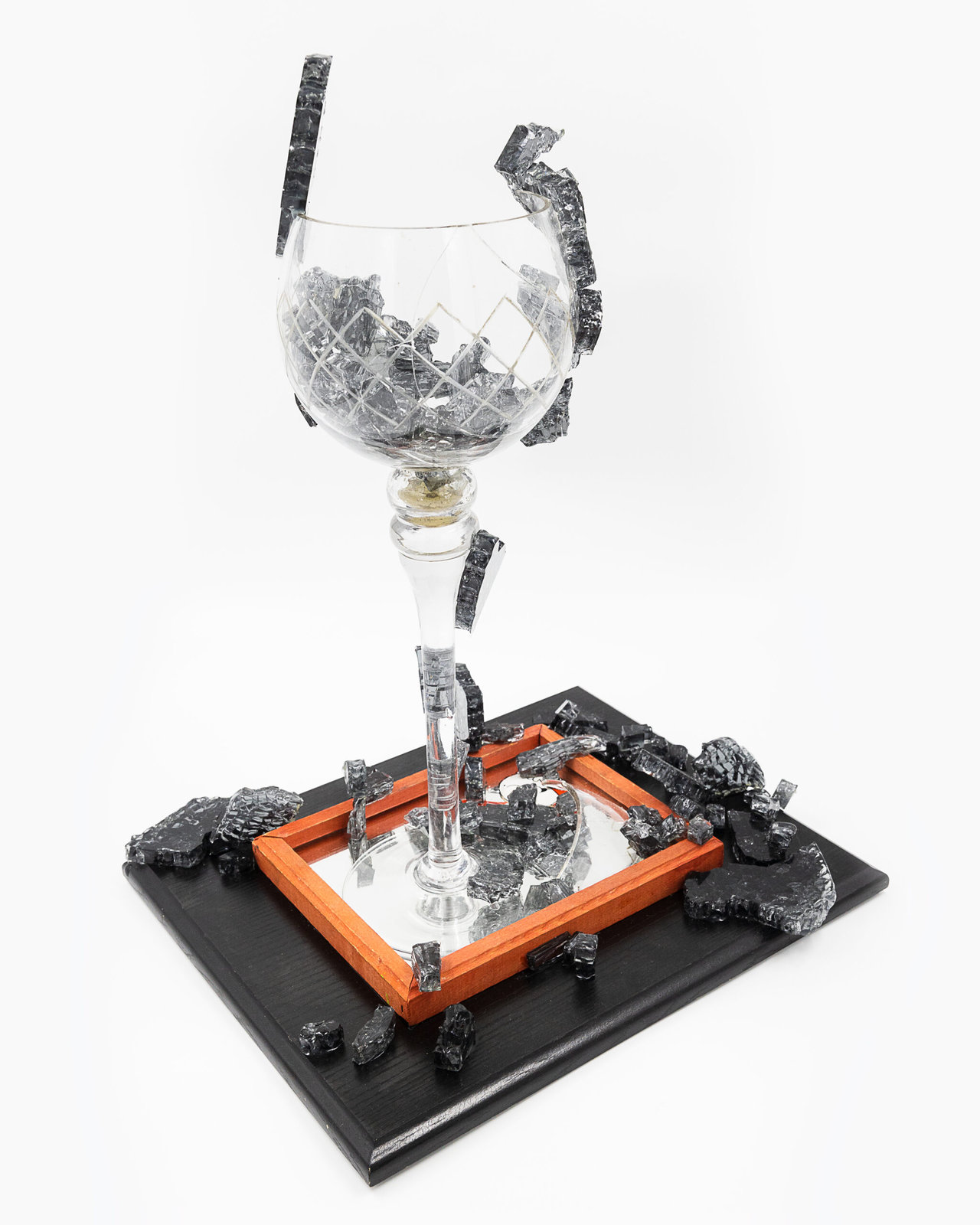 Tall candle holder (without the candle) in the shape of a champagne glass, is broken and reconstructed. Chunks of black shattered glass appear to burst out of it. The champagne glass stands on a red wood-framed mirror made by Tom Wilson (tm) and pieces of shattered black glass are strewn about. All this sits on a black wooden base.